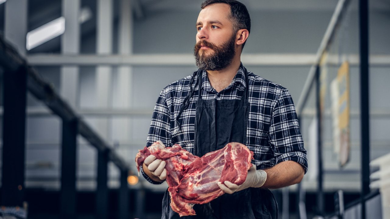 Portrait of a bearded meat man dressed in a fleece shirt holds fresh cut meat in a market.